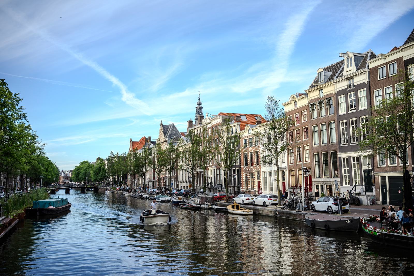 A tree-lined Amsterdam canal with moored boats and tall gabled houses under a blue sky