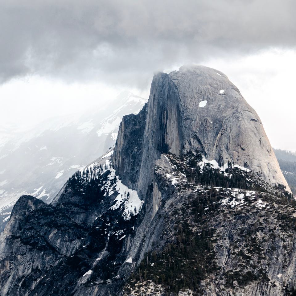 The sheer face of Half Dome rising above snowy, forested ridges, its top lost in low storm cloud