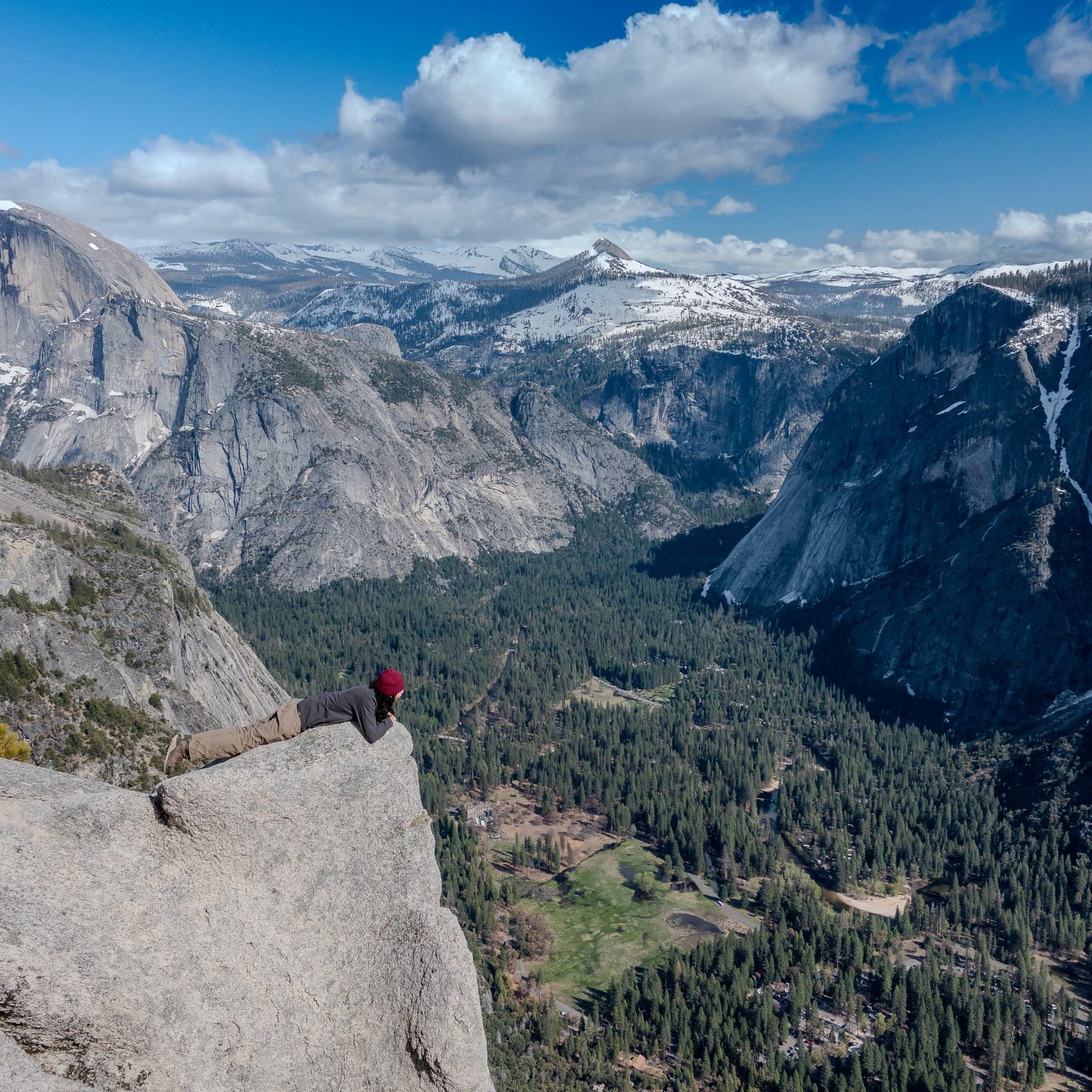 A person lying on the edge of a granite ledge high above Yosemite Valley, with snowy peaks and sheer cliffs beyond
