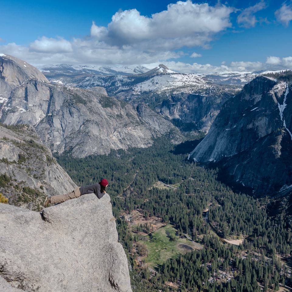 A person lying on the edge of a granite ledge high above Yosemite Valley, with snowy peaks and sheer cliffs beyond